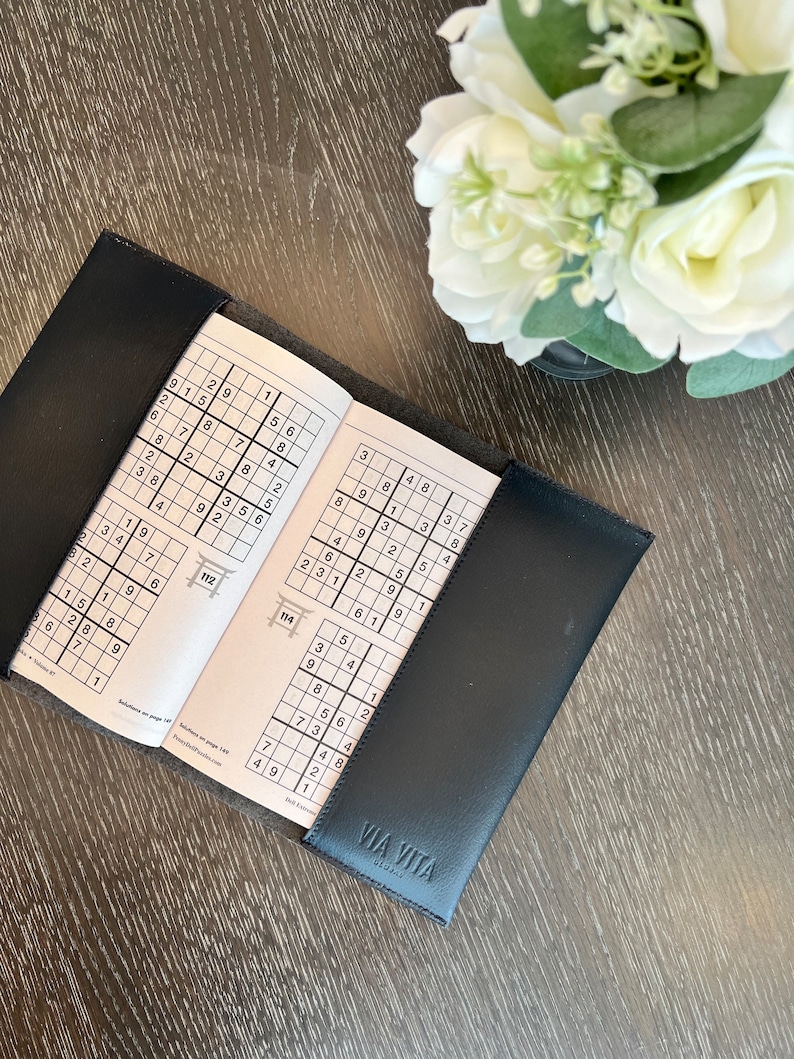 May include: An open black leather-bound book with a Sudoku puzzle on each page. The book is open to page 112 and 114. The text "VIA VITA" is printed on the cover.