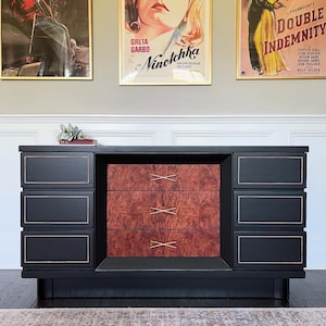 May include: A black mid-century modern dresser with gold trim and a burl wood center. The dresser has six drawers and is set against a wall with vintage movie posters, including "Ninotchka" and "Double Indemnity."
