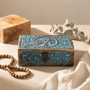 Peut inclure: Une boîte rectangulaire en bois avec un motif d'arbre de vie sculpté à la main sur le couvercle. La boîte présente une finition vieillie avec des tons de bois naturel brun et des accents de peinture bleue vive. Un loquet en métal sombre est fixé.
