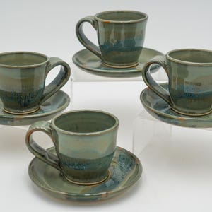 May include: Set of four green and blue ceramic teacups and saucers. The cups and saucers have a mottled glaze with a light green base and darker blue and green streaks.