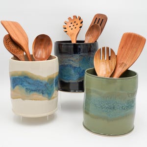 May include: Three ceramic utensil holders in different colors, each with a unique glaze pattern. The holders are filled with wooden kitchen utensils, including spoons, spatulas, and a slotted spoon.