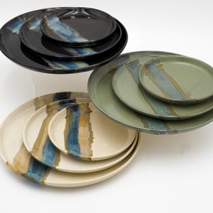May include: A selection of ceramic plates in different sizes and colours. The plates have a distinctive design with blue, tan, and green stripes. The plates are stacked and arranged on a white surface, highlighting their artistic appeal.