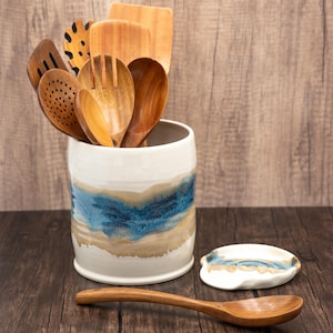 May include: A white ceramic utensil holder with a blue and beige glaze design. The holder is filled with wooden spoons and spatulas.