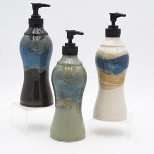 Könnte beinhalten: Drei Keramik-Seifenspender mit schwarzen Pumpköpfen. Die Spender haben eine einzigartige, geschwungene Form und sind mit einer Mischung aus Farben bemalt, darunter Blau, Braun und Creme. Sie werden auf transparenten Acrylständern ausgestellt.