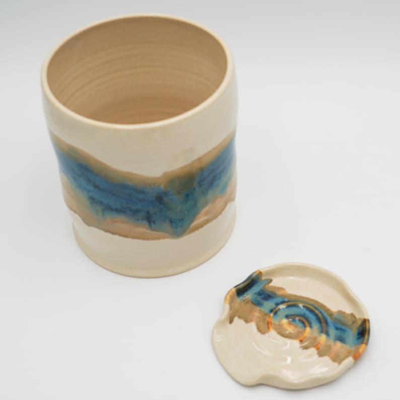 May include: A ceramic jar with a lid, both featuring a blue and brown glaze pattern. The jar is white with a wide mouth and a rounded shape. The lid is a smaller, round dish with a slight dip in the center.