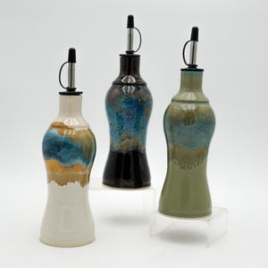 May include: Three ceramic oil bottles with stainless steel spouts. The bottles have a unique shape and are glazed in various colors, including white, blue, black, and green. Each bottle is displayed on a clear acrylic stand.