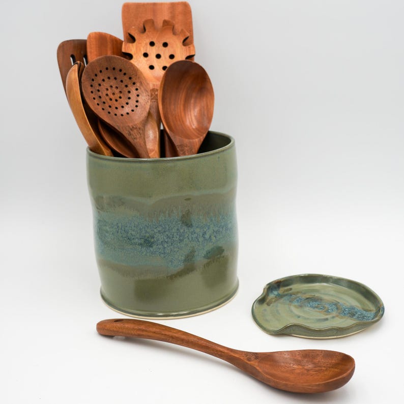 May include: A green ceramic utensil holder with a blue and green glaze. The holder is filled with wooden spoons and spatulas. A matching green ceramic spoon rest is also shown.