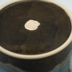 May include: A dark brown ceramic pot with a light brown rim and a blue and grey glaze. The bottom of the pot has a label that reads "John Green Pottery".