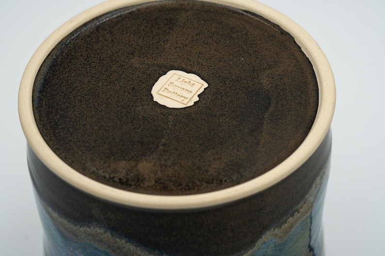 May include: A dark brown ceramic pot with a light brown rim and a blue and grey glaze. The bottom of the pot has a label that reads "John Green Pottery".