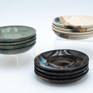 May include: A stack of six ceramic saucers in shades of green, blue, and brown. The saucers are stacked on a clear acrylic stand. Another stack of six saucers in shades of white, brown, and blue are stacked on a clear acrylic stand.
