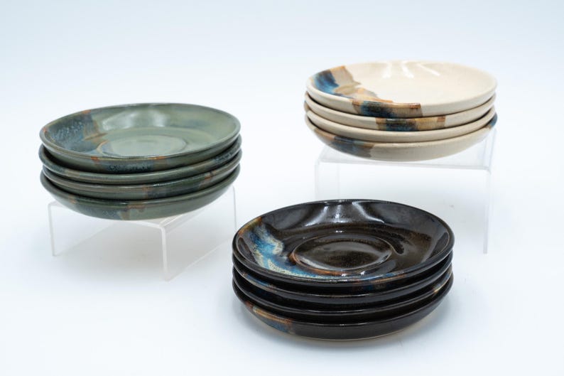 May include: A stack of six ceramic saucers in shades of green, blue, and brown. The saucers are stacked on a clear acrylic stand. Another stack of six saucers in shades of white, brown, and blue are stacked on a clear acrylic stand.