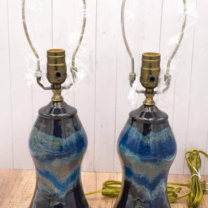 May include: Two ceramic table lamps with a dark blue and teal glaze. The lamps have a bulb socket and a brass-colored neck. The lampshades are not included. The lamps are on a wooden surface.
