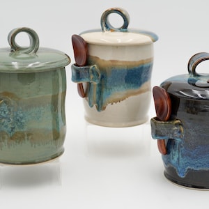 Puede incluir: Tres tarros de cerámica con tapas y cucharas de madera adjuntas. Los tarros están esmaltados en tonos de verde, azul y negro. Cada tarro tiene un asa de bucle en la tapa y un pequeño soporte para la cuchara. Los tarros miden aproximadamente 13 cm de alto.