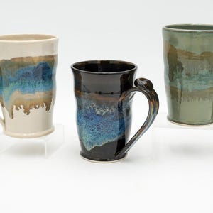 Puede incluir: Tres tazas de cerámica con asas únicas y diseños de esmalte coloridos. Las tazas presentan un color base crema, negro y verde, con acentos azules, marrones y verdes. Cada taza mide aproximadamente 13 cm de alto.