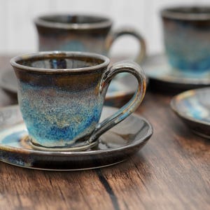 May include: Set of four ceramic coffee cups and saucers with a blue and brown glaze. The cups have a rounded shape and a handle. The saucers are round and have a slight lip.
