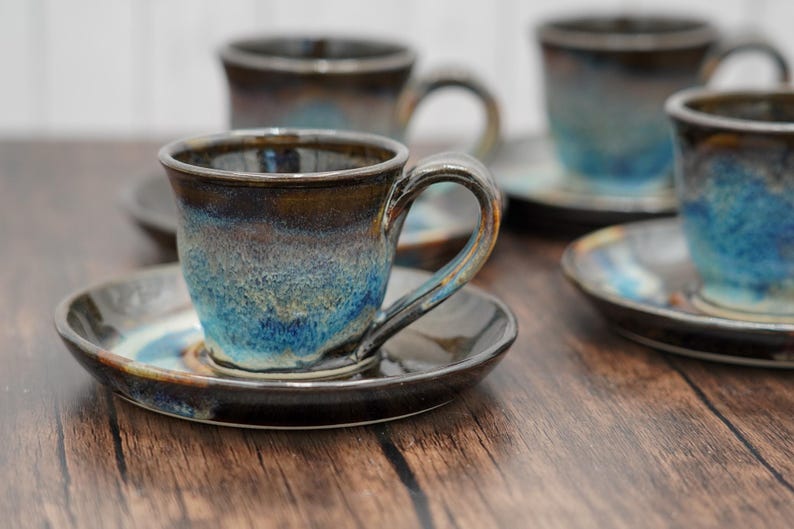 May include: Set of four ceramic coffee cups and saucers with a blue and brown glaze. The cups have a rounded shape and a handle. The saucers are round and have a slight lip.