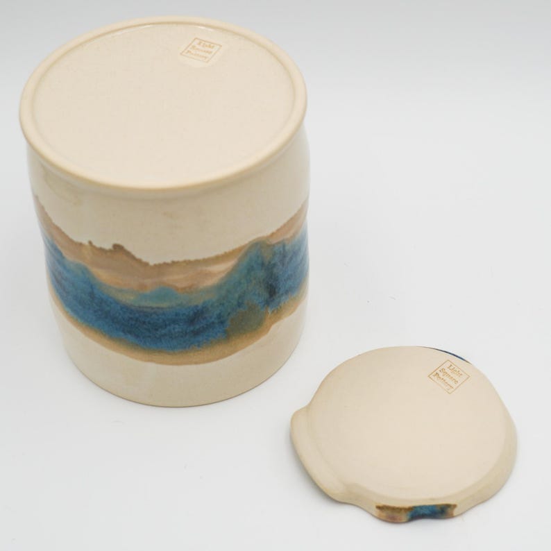 May include: A white ceramic jar with a blue and brown glaze design. The jar has a lid and is handmade. The lid has a small stamp that reads "Made in America".