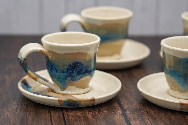 May include: Set of four ceramic coffee cups and saucers with a blue, brown, and white glaze. The cups have a rounded shape and a handle.