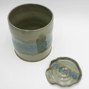 May include: A green ceramic jar with a lid. The jar has a blue and brown glaze with a speckled finish. The lid has a wavy edge and a similar glaze.
