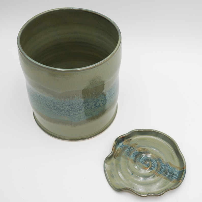 May include: A green ceramic jar with a lid. The jar has a blue and brown glaze with a speckled finish. The lid has a wavy edge and a similar glaze.