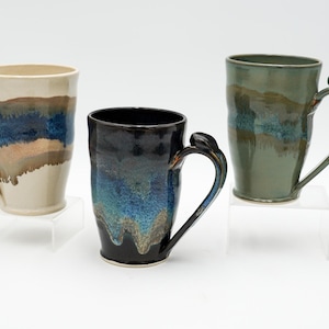 May include: Three ceramic mugs with distinctive handles and colourful glazes. The mugs are in shades of cream, blue, brown, and green. Each mug sits on a clear acrylic stand, showcasing the handcrafted details and artistic design.