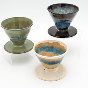 May include: Three ceramic pour-over coffee drippers in shades of green, blue, brown, and cream. Each dripper has a conical shape and sits on a circular base. The drippers are displayed on clear acrylic stands against a white background.