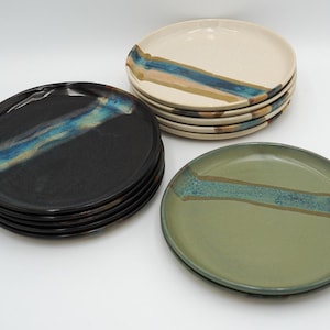 May include: A collection of ceramic plates in various colors. There are stacks of black, cream, and green plates. Each plate has a diagonal stripe of blue, teal, and brown glaze. The plates are round and appear to be handmade.