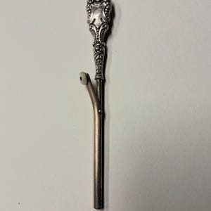 May include: A silver-toned metal tool featuring an ornate, embossed handle with intricate scrollwork and a shield-like design at the top. The shaft includes a hinged, angled arm extending outward, tipped with a small, circular white plastic bead.