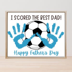 以下が含まれることがあります： サッカーボールと青い手形が描かれたフレームプリント。テキストは「I SCORED THE BEST DAD！」と「Happy Father's Day」です。フレームは明るい木の色です。父の日のプレゼントに最適です。