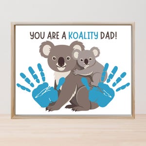 Puede incluir: Impresión enmarcada con una ilustración de un koala y un bebé koala con huellas de manos azules. El texto "YOU ARE A KOALITY DAD!" se muestra en azul y negro. La obra de arte está sobre un fondo blanco.