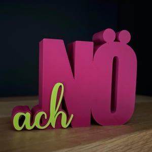 Könnte beinhalten: Ein magentafarbenes Holzschild mit dem Wort "Nö ach" in einer verspielten Schriftart. Das Wort "Nö" ist in fetten Blockbuchstaben geschrieben, und das Wort "ach" in einer kursiven, limettengrünen Schriftart. Das Schild steht auf einem Holzregal.