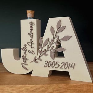 Könnte beinhalten: Dekorative Holzskulptur in Form der Buchstaben "JA" mit den Namen "Natalie & Andreas" und dem Datum "3005-2014" eingraviert. Das "A" ist mit einem Blattdesign verziert. Auf dem "J" befindet sich ein kleines Glasfläschchen mit einem Korken.