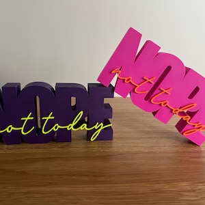 Könnte beinhalten: Zwei 3D-gedruckte Wortkunstwerke. Eines ist lila und liest "NOPE" mit "not today" in gelber Schrift. Das andere ist rosa und liest "NOPE" mit "not today" in orangefarbener Schrift. Beide stehen auf einer Holzoberfläche.