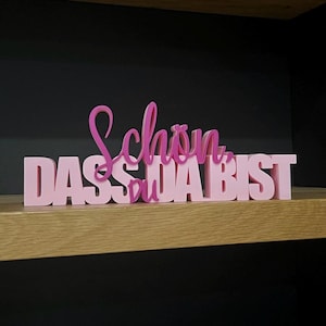 Könnte beinhalten: Ein rosa Dekoschild mit den Worten "Schön dass du da bist" in einem geschichteten Design. Das Schild wird auf einem Holzregal vor dunklem Hintergrund ausgestellt. Der Text ist in einer Schreibschrift.