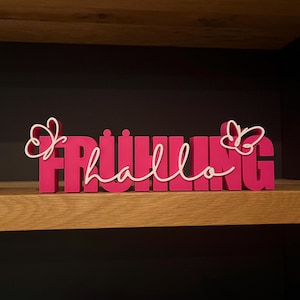 Könnte beinhalten: Ein leuchtend pinkfarbenes Holzschild mit der Aufschrift "FRÜHLING hallo" in einer kräftigen, blockartigen Schrift, mit weißer Schreibschrift und Schmetterlingsakzenten. Das Schild steht auf einem Holzregal vor dunklem Hintergrund.
