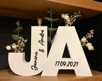 May include: A white wooden sign with the letters "JA", the names "Janina & Andre", and the date "17.09.2021" in black. The letters are decorated with small floral arrangements and a miniature glass bottle.