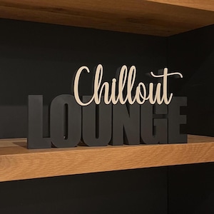 Könnte beinhalten: Ein Holzschild mit der Aufschrift "Chillout LOUNGE". Das Wort "Chillout" ist in Schreibschrift geschrieben, während "LOUNGE" in Blockbuchstaben gehalten ist. Das Schild steht vor einem schwarzen Hintergrund und liegt auf einem Holzregal.