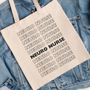 May include: A beige tote bag with the words "NEURO NURSE" printed in black. The text is repeated multiple times in a diagonal pattern. The bag is placed on a light blue denim jacket.
