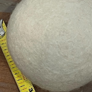 May include: A round, off-white felted wool ball positioned next to a yellow measuring tape. The tape shows the diameter of the sphere is approximately 3 inches. The surface of the ball has a textured, fibrous appearance consistent with felted wool.