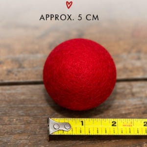 May include: A solid red felted wool ball with a textured surface. The text "APPROX. 5 CM" is printed above the ball, and a yellow measuring tape is positioned at the base, showing a diameter of approximately 2 inches for the round object.