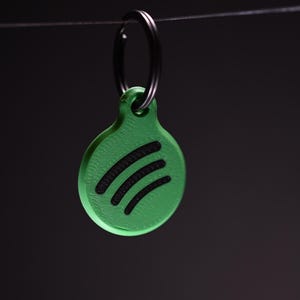 May include: A green keychain with a black key ring. The keychain is a rounded shape with a green background and three black curved lines. The background is a dark gray.