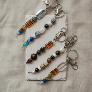 Peut inclure: Quatre porte-clés perlés avec des fermoirs argentés. Chaque porte-clés présente une combinaison unique de perles de verre colorées, notamment bleues, oranges, blanches et noires. Les porte-clés sont disposés sur une surface texturée blanc cassé.