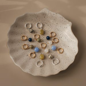 May include: A selection of hoop earrings featuring a variety of gemstone beads in shades of blue, green, white, and speckled patterns. The earrings are arranged on a speckled, off-white ceramic plate. Some hoops are gold, others silver.