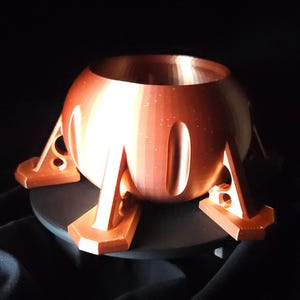 Może przedstawiać: Miedziany, kulisty obiekt z szerokim otworem i czterema trójkątnymi nogami. Nogi mają okrągłe wycięcia i są przymocowane do okrągłej podstawy. Obiekt jest ustawiony na ciemnym tle.