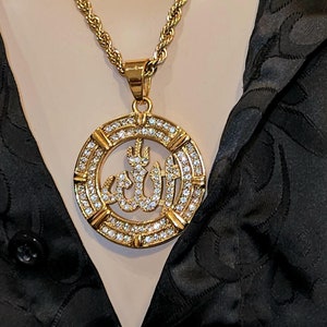 Puede incluir: Un collar de cadena de cuerda dorada con un colgante que presenta la palabra árabe para Dios, "Allah", rodeado de brillantes diamantes de imitación transparentes. El colgante es circular y tiene un marco dorado.