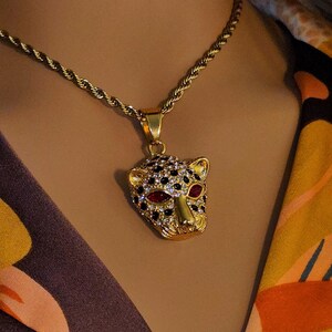 Puede incluir: Un collar de cadena de oro con un colgante de oro en forma de cabeza de leopardo. El colgante está incrustado con diamantes de imitación negros y transparentes, y los ojos son rojos.