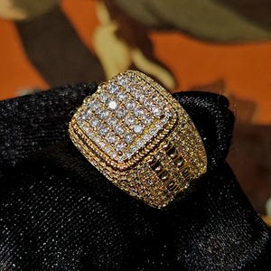 May include: A gold-colored ring with a square face, encrusted with numerous small, sparkling gemstones. The ring has a detailed, textured band and is set against a dark background, showcasing its brilliance.