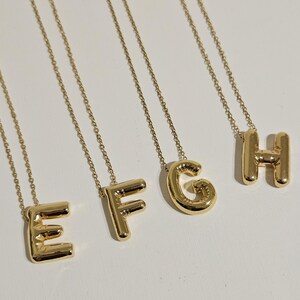 May include: Four gold-colored initial necklaces, spelling out the letters E, F, G, and H. Each letter is a bold, rounded font, suspended from a delicate chain. The necklaces are displayed against a plain white background, highlighting the jewelry.