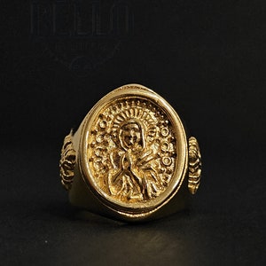 Puede incluir: Un anillo de oro con un diseño ovalado que presenta una imagen detallada de una persona con alas. El anillo tiene una banda texturizada con un diseño decorativo a cada lado.