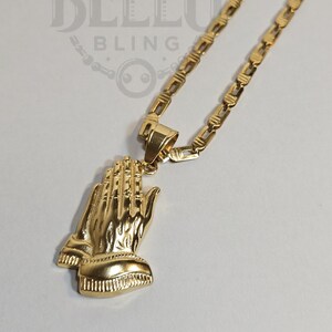 Puede incluir: Un collar de cadena dorada con un colgante de manos en oración. La cadena es de estilo curb.